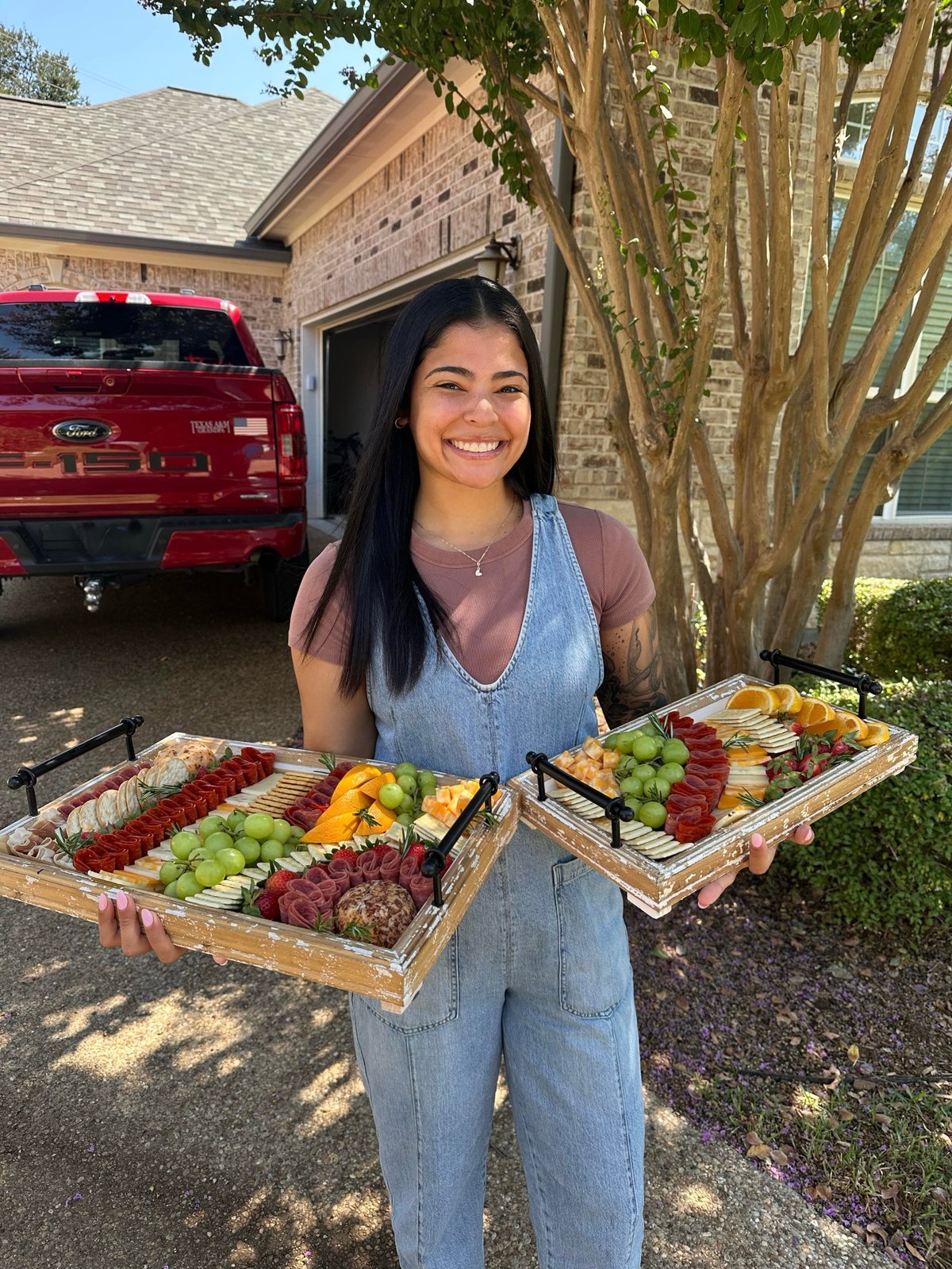 Woman smiling while holding wooden charcuterie boards with fresh fruits, meats, and cheeses in a driveway